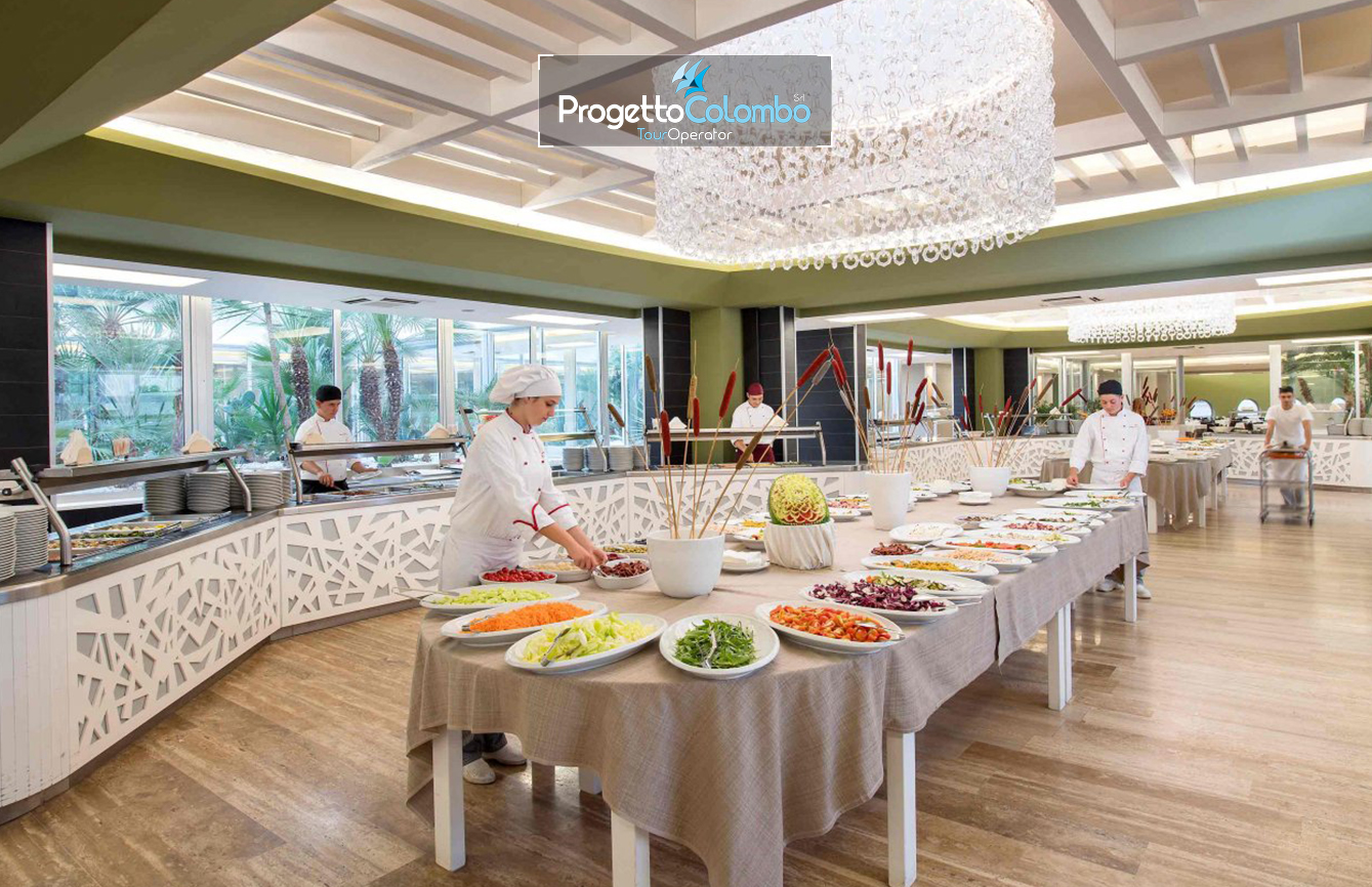 Progetto Colombo Bari, Tour Operator, Mare Italia, Voi Alimini resort, Alpitour, Bravo Club, All Inclusive, Top Club, Puglia, Salento_Buffet 2 Spiaggia e Mare: "Spiaggia di sabbia bianca e mare cristallino del VOI Alimini Resort a Otranto, ideale per vacanze a metà settembre." Piscina: "Ampia piscina all'aperto circondata dal verde del giardino nel resort VOI Alimini in Puglia." Ristorazione: "Buffet vario e colorato nella sala ristorante del VOI Alimini Resort per gli ospiti in formula all-inclusive." Organizzazione Progetto Colombo Viaggi Bari